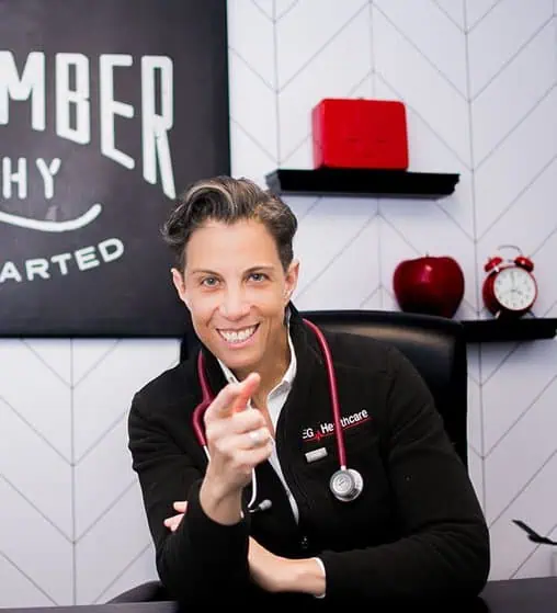 Person wearing a black jacket with a stethoscope around their neck is sitting at a desk in a medical office, pointing towards the camera. There is a sign, two shelves, and several items on the wall behind them.